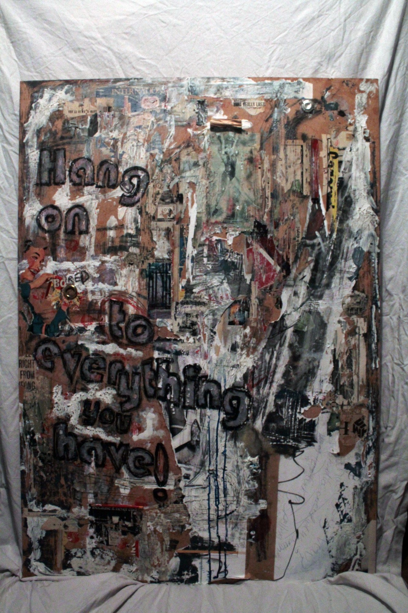 Full view of a hanging sculptural collage with layered advertising imagery, torn paper and large black lettering.