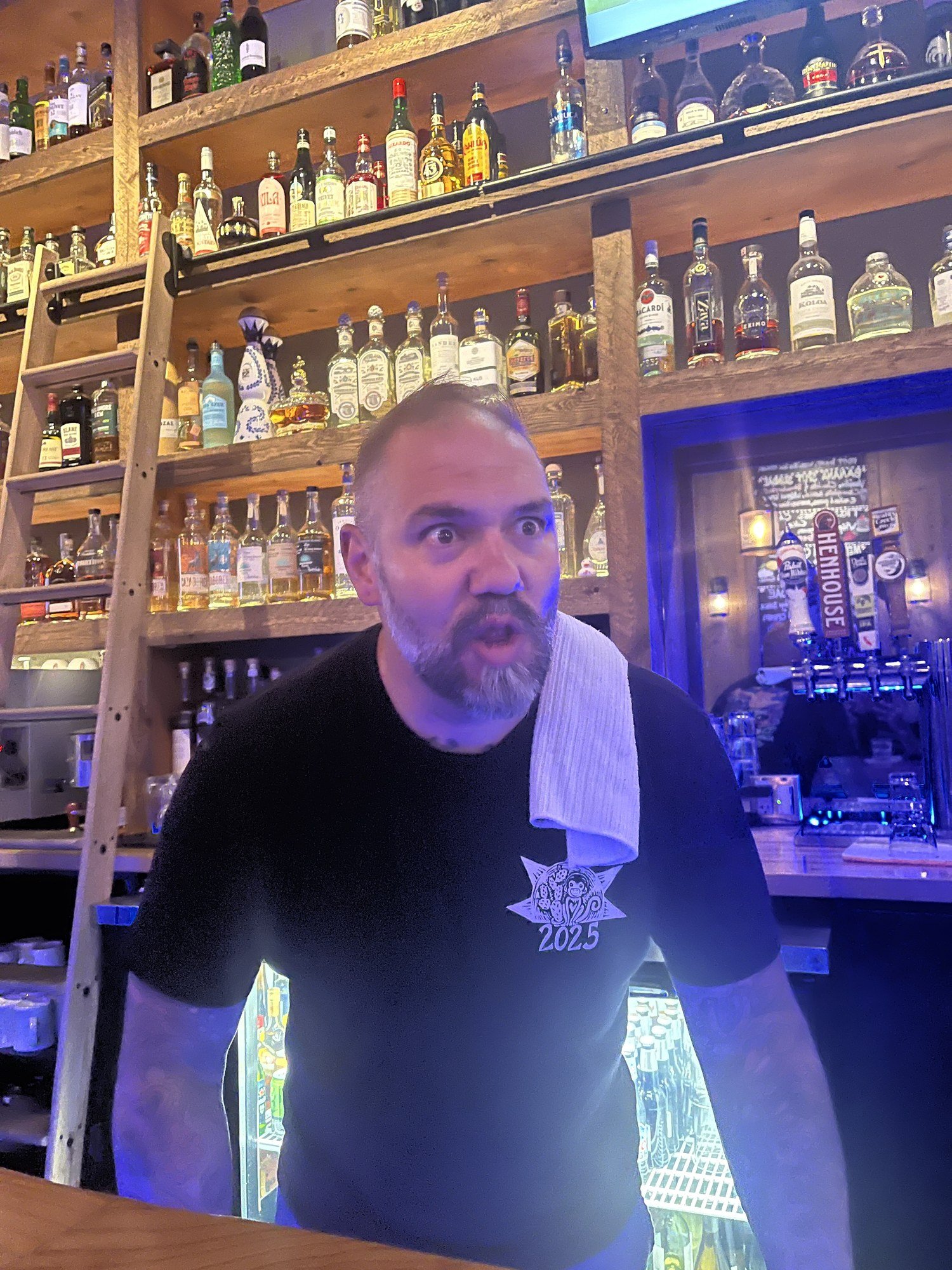 A smiling man stands behind the bar with a towel over one shoulder.