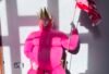 A seated performer in a vivid pink costume and crown holds a distressed American flag in a sunlit interior.