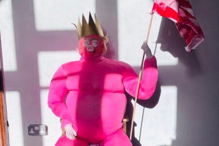 A seated performer in a vivid pink costume and crown holds a distressed American flag in a sunlit interior.